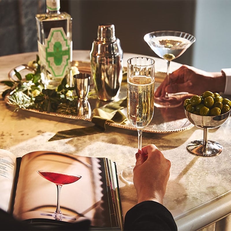A festive drinks scene with champagne, martinis, olives, a cocktail shaker, and an open cocktail book on a marble table.