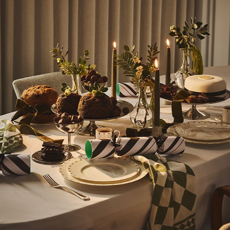 A Christmas dining table set with puddings, a white-iced cake, chocolates, candles, and crackers, decorated with greenery.