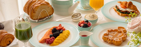 Breakfast table set with sliced tropical fruit, pastries, bread, a croissant sandwich, green juice, orange juice, tea, and a waffle on pale blue and white china.