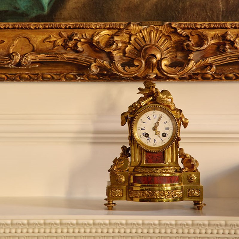 Ornate gold mantel clock with Roman numerals, placed below a gilded picture frame.