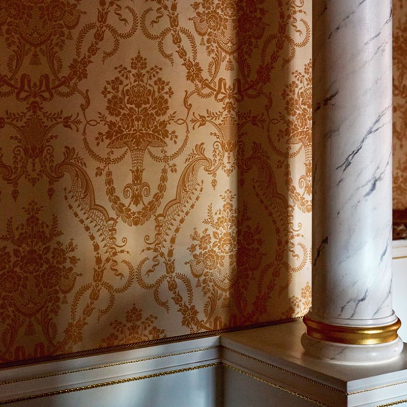 Gold damask-patterned wallpaper next to a white marble column with gold trim.