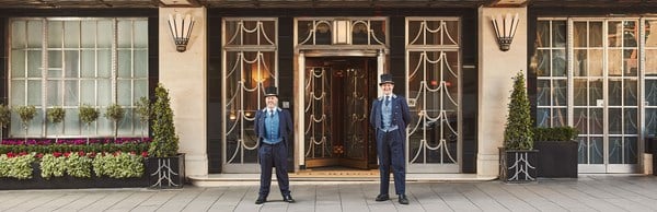 Wide view of Claridge’s entrance with two doormen in blue uniforms framed by Art Deco windows and potted plants.