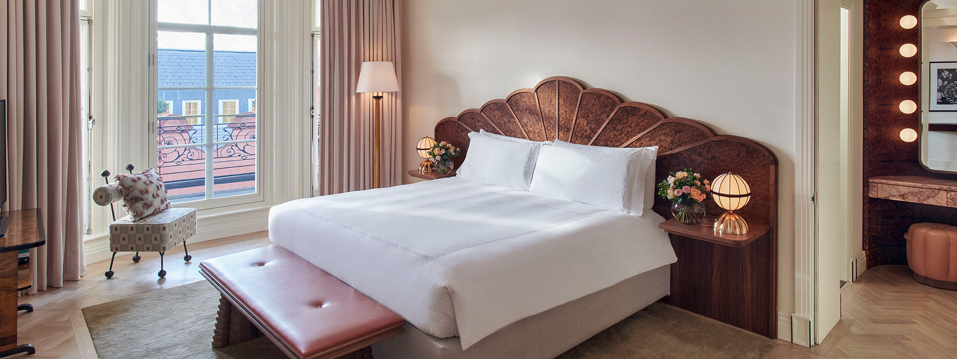 A bed in an Art Deco atmosphere, surrounded by shades of pink, with an armchair by the window.