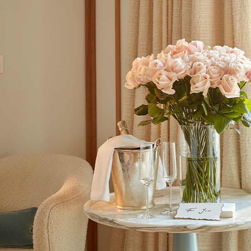 A refined setting featuring a grand bouquet of blush pink roses, a chilled champagne bucket, two crystal glasses, and a delicate handwritten note, enhancing the scene’s romantic allure.