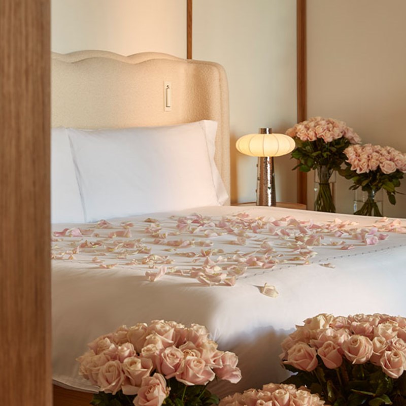 A hotel bed dressed in crisp white linens, scattered with pink rose petals, with bouquets of fresh roses placed on the bedside tables, creating an intimate and luxurious retreat.