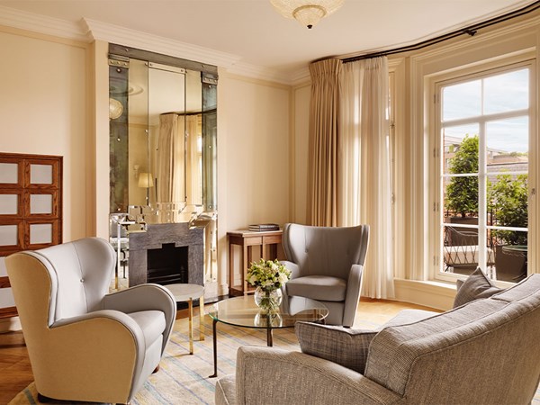 Bright lounge with grey armchairs and sofa around a glass table, with tall windows opening to a terrace.